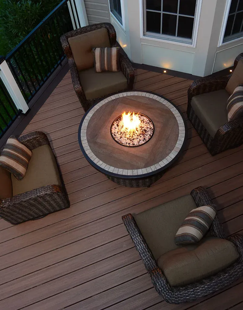 Outdoor deck with a fire pit surrounded by four wicker chairs with pillows.