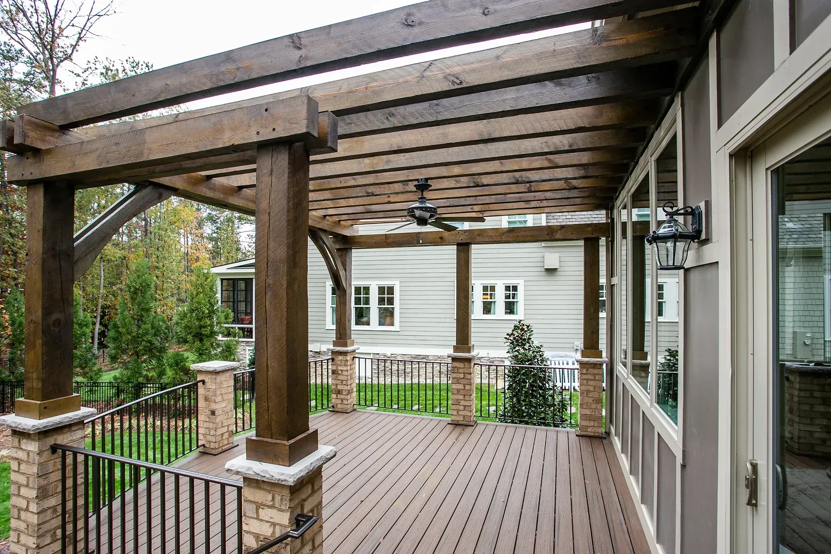 Wooden pergola with brown decking, supported by stone and wood pillars, overlooking a yard.