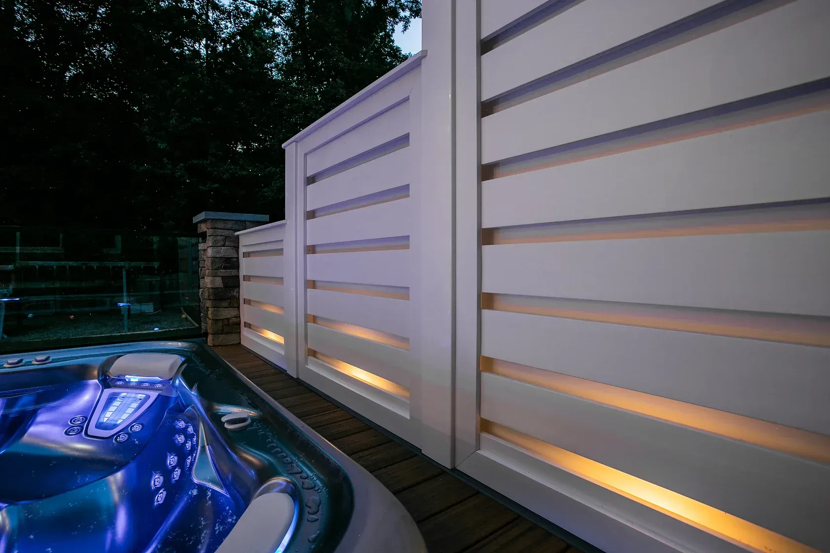 Hot tub with white slatted fence and built-in lighting on a wooden deck at night.
