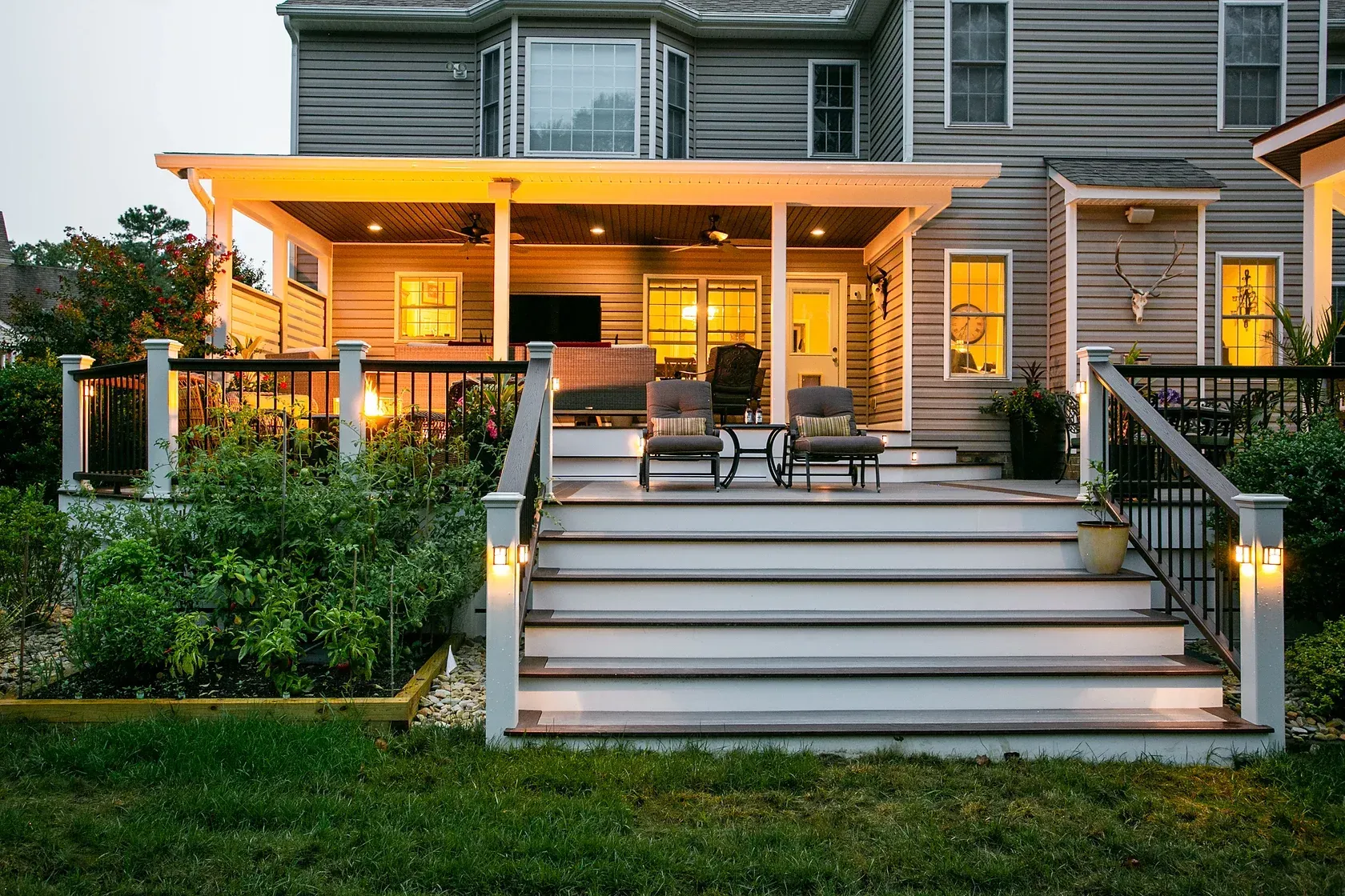 Backyard deck with steps, seating, and overhead porch, lit with warm lights at dusk.