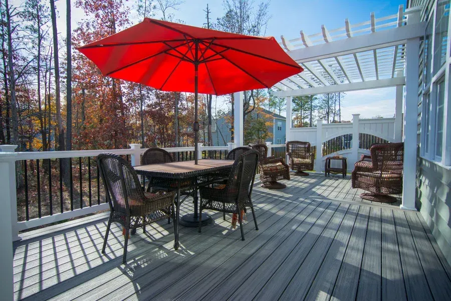 Patio with table, chairs, red umbrella, and pergola.  Deck overlooks trees and blue sky.