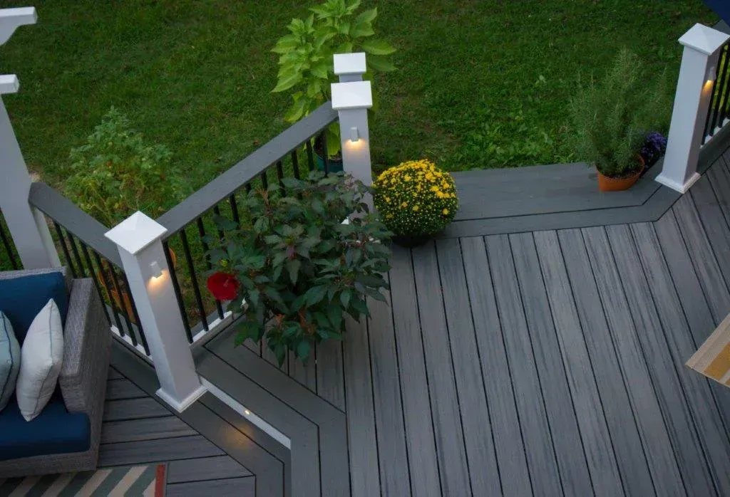 Overhead view of a gray deck with potted plants and dark railing. Green lawn and blue chair visible.