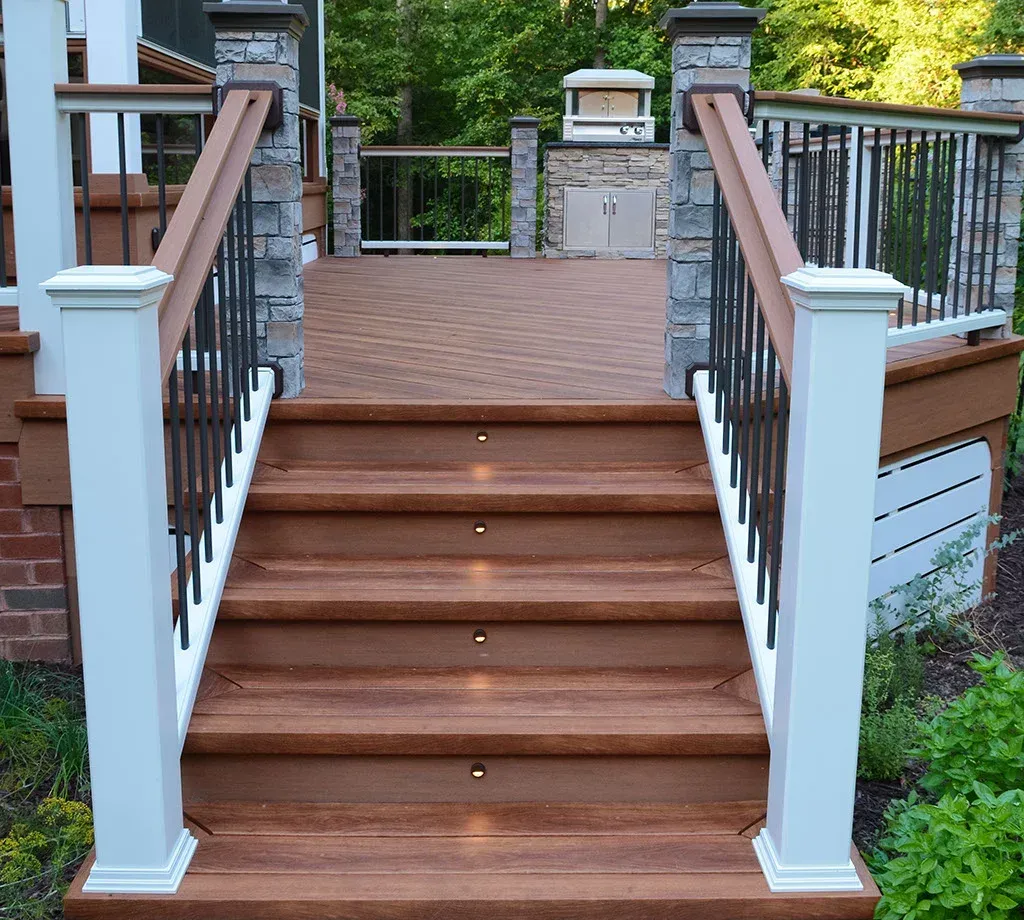 Wooden deck stairs with built-in lights, flanked by white posts, stone accents, and black metal railings.