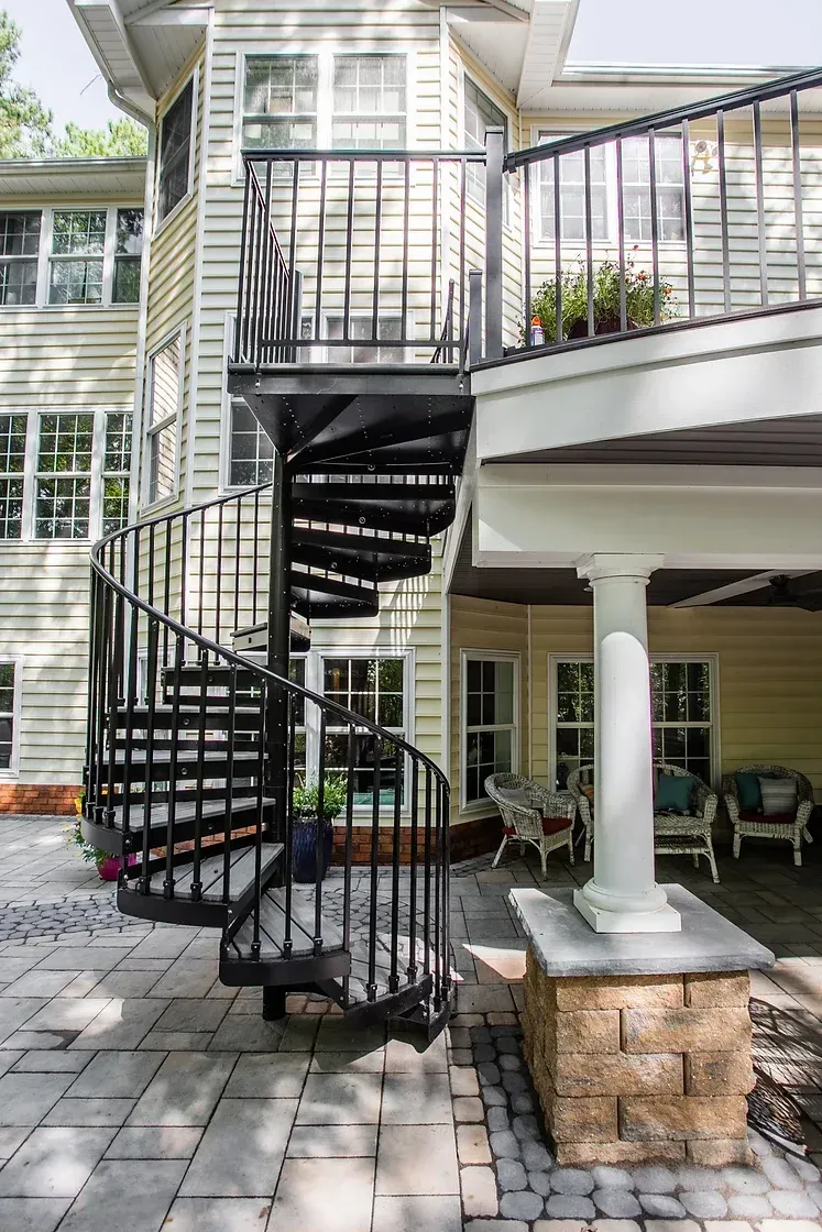 Black spiral staircase on a patio, leading to a balcony, beside a yellow house.