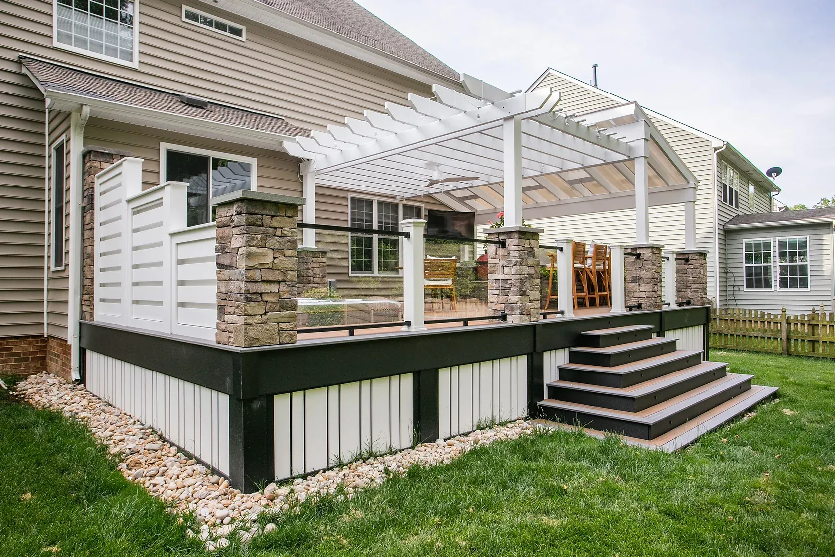 Deck with pergola attached to a beige house, with stone columns, white railing, and stairs.