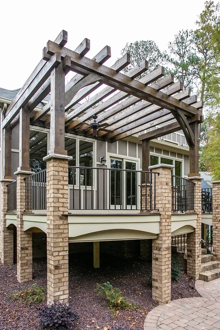 Wooden pergola over a raised deck with a light brown siding building, brick columns, and a stone walkway.