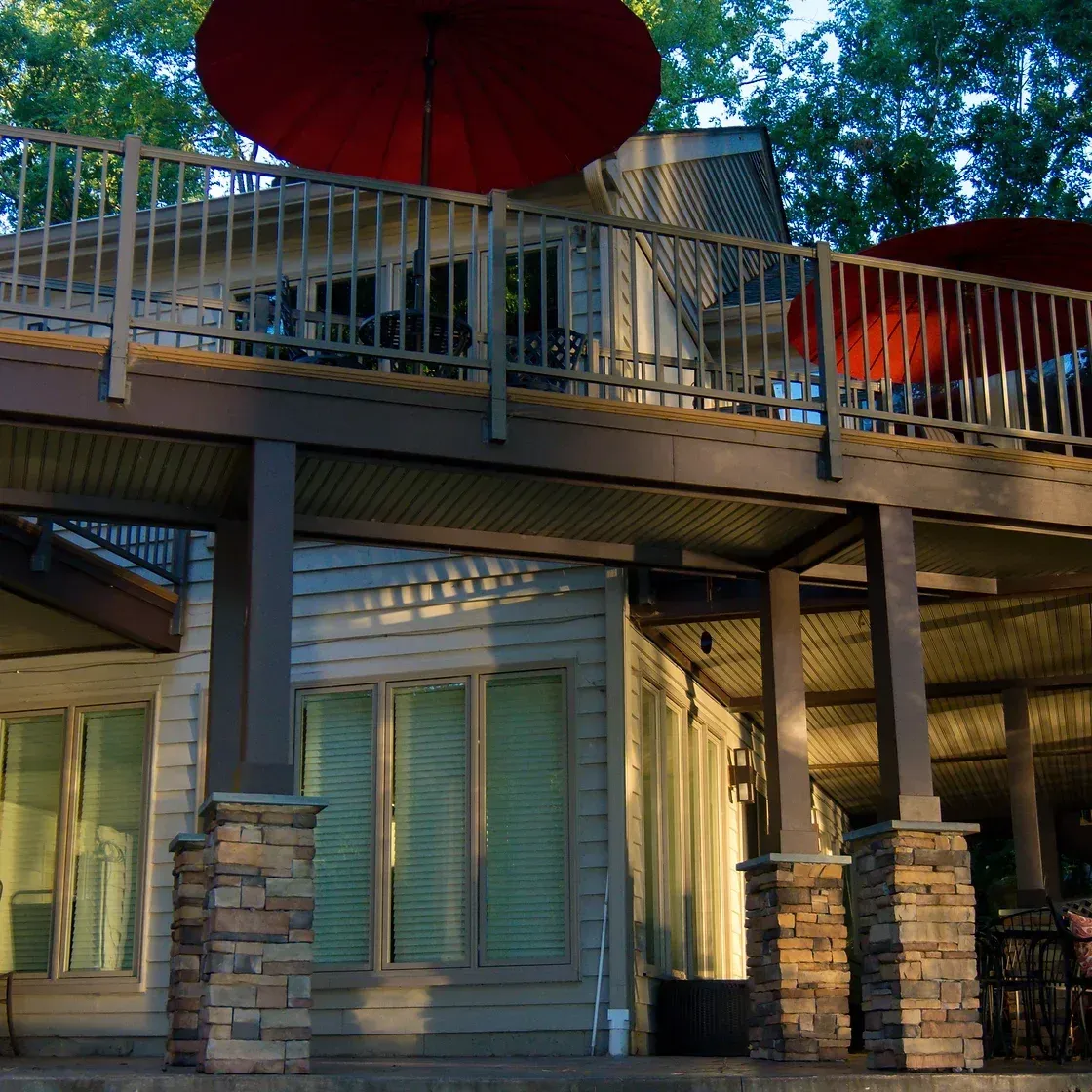 Two-story house with a deck. Red umbrellas, stone columns, and windows are visible.