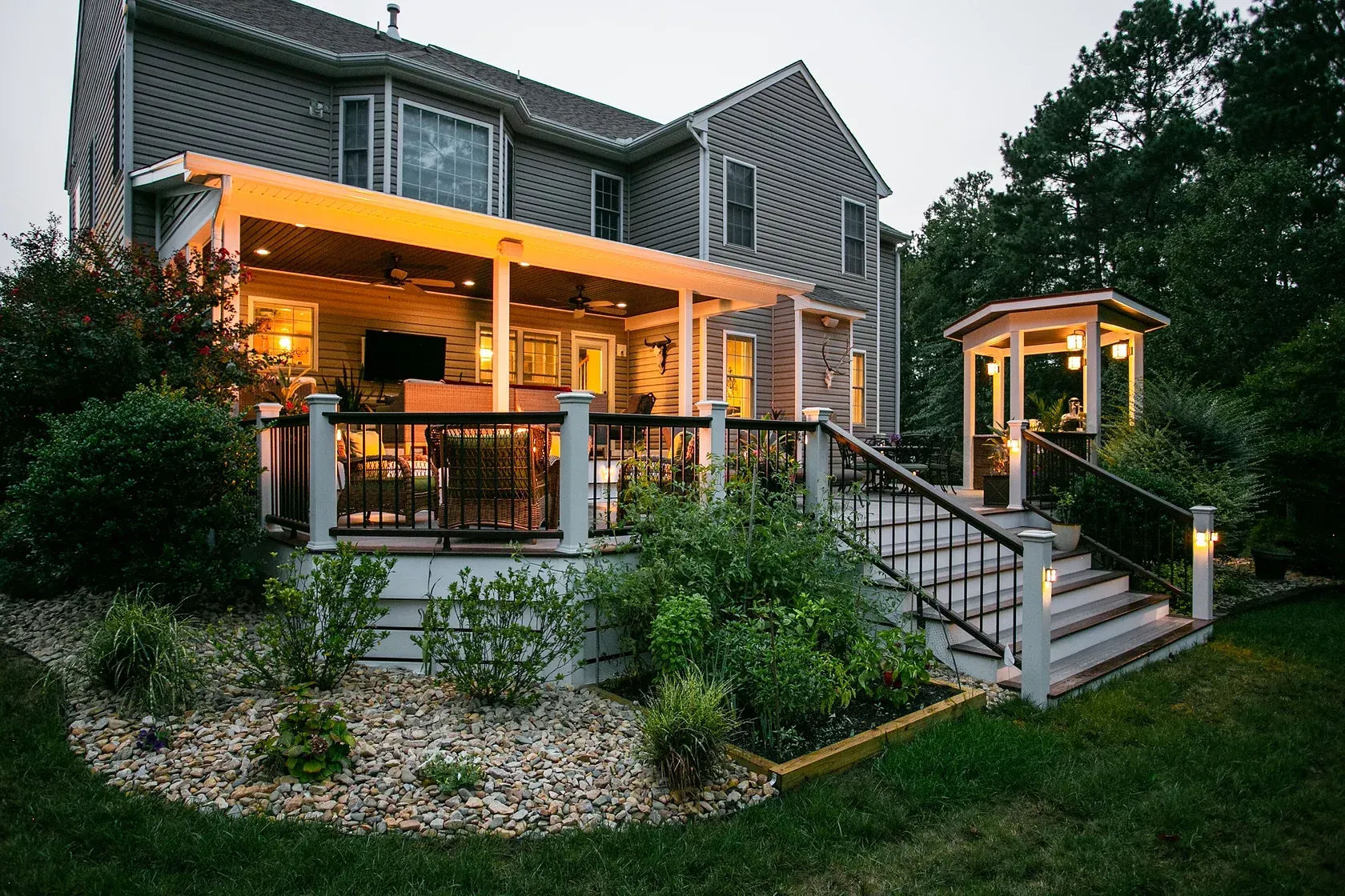 Backyard deck with pergola, stairs, and gazebo, lit at dusk. Landscaping and a large house in the background.