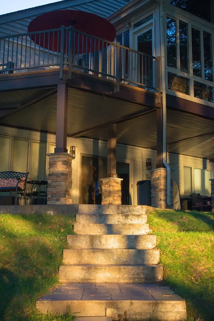 Stone steps lead up to a deck with an umbrella and a house with large windows.