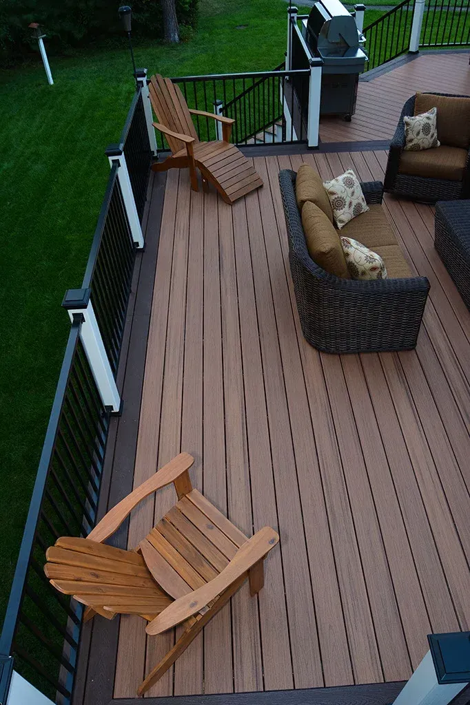 Wooden deck with Adirondack chairs, wicker sofa, and grill, overlooking green lawn.