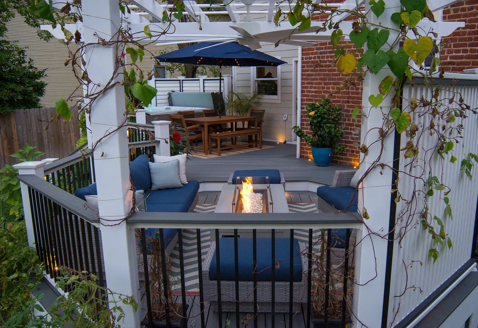 Outdoor deck with seating, fire pit, pergola, and climbing vines. Blue cushions, gray deck.