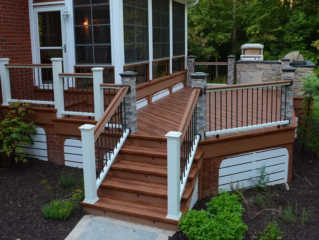 Wooden deck with ramp and steps, connected to a screened porch, surrounded by landscaping.