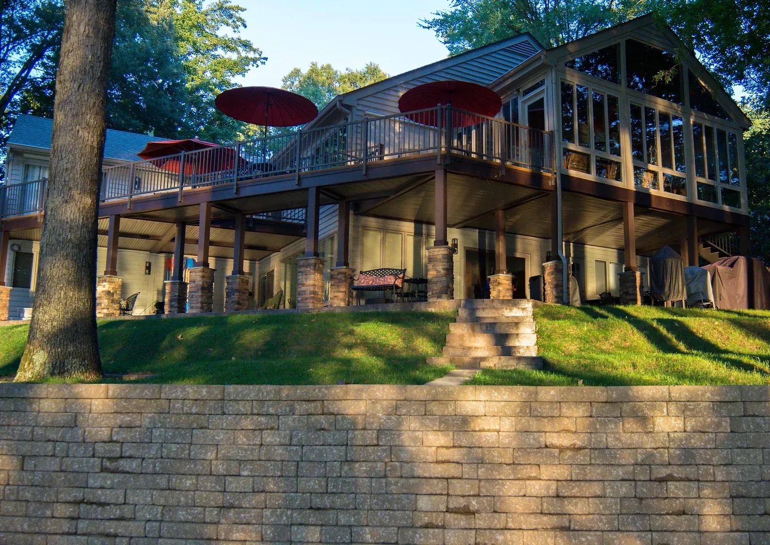 Two-story lakeside home with elevated deck, brick wall, and red umbrellas.