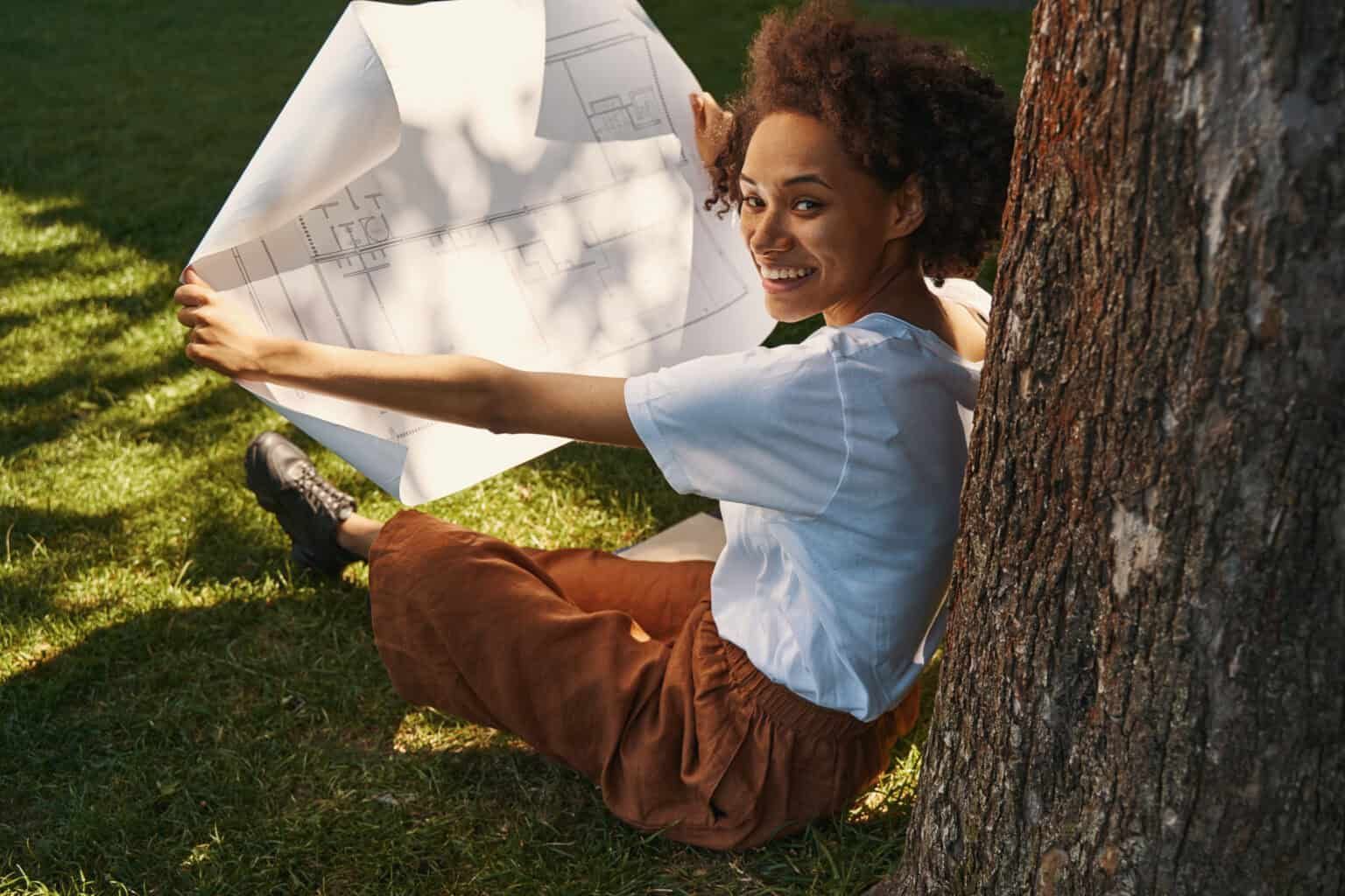 Woman seated on grass by a tree, unfurling blueprints, smiling.