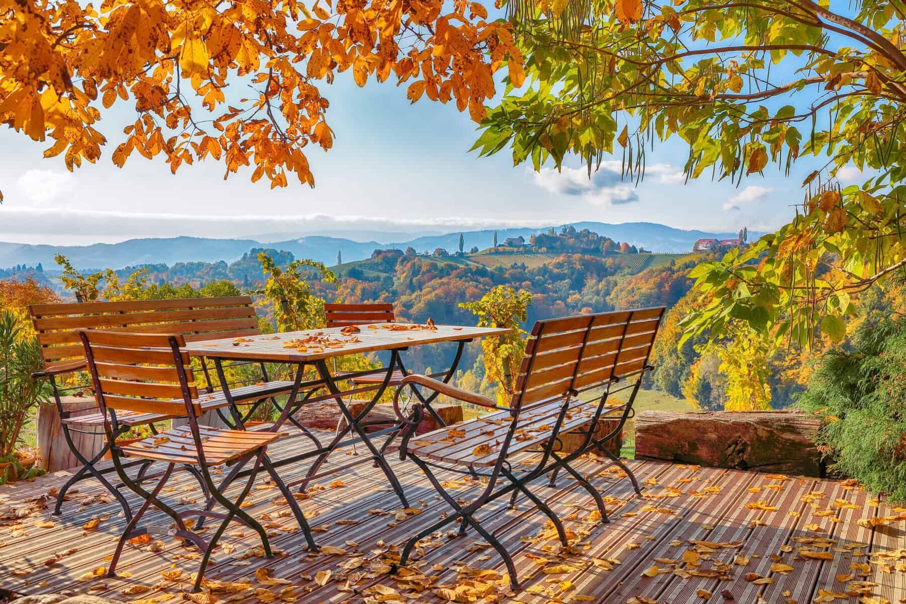 Wooden table and chairs on a deck, autumn foliage, scenic view of hills and trees under a blue sky.