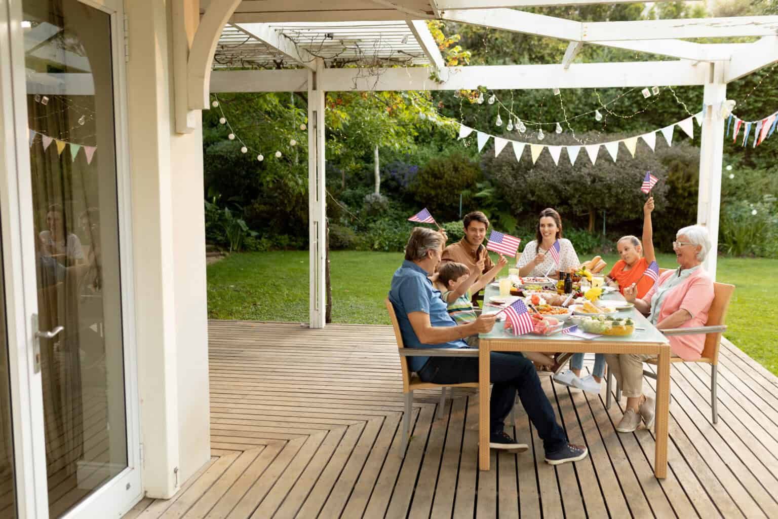 Family celebrating outdoors, gathered at a table decorated with flags, under a pergola, on a deck.