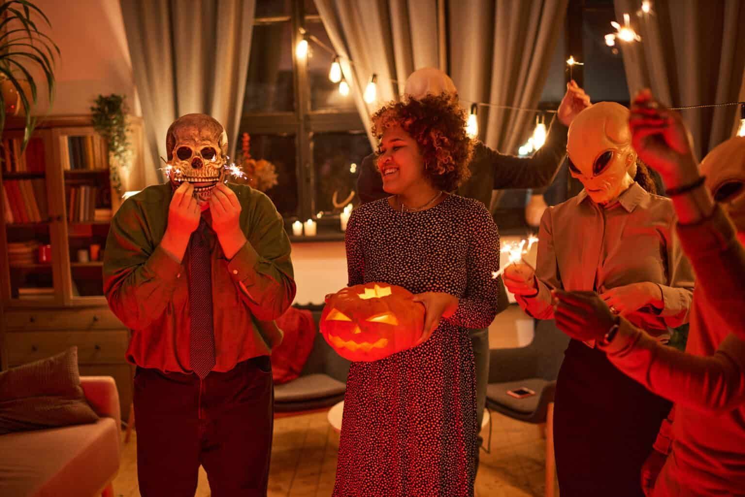 People in costumes at a Halloween party, holding a skull, a carved pumpkin, and sparklers in a warmly lit living room.