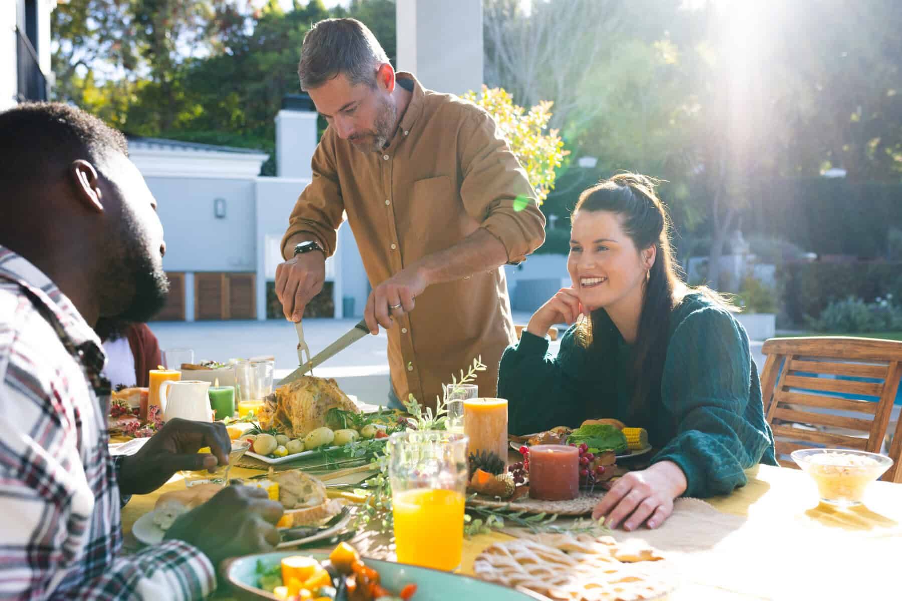 Man carving a roasted chicken at an outdoor table; two people smile. Sunlight.