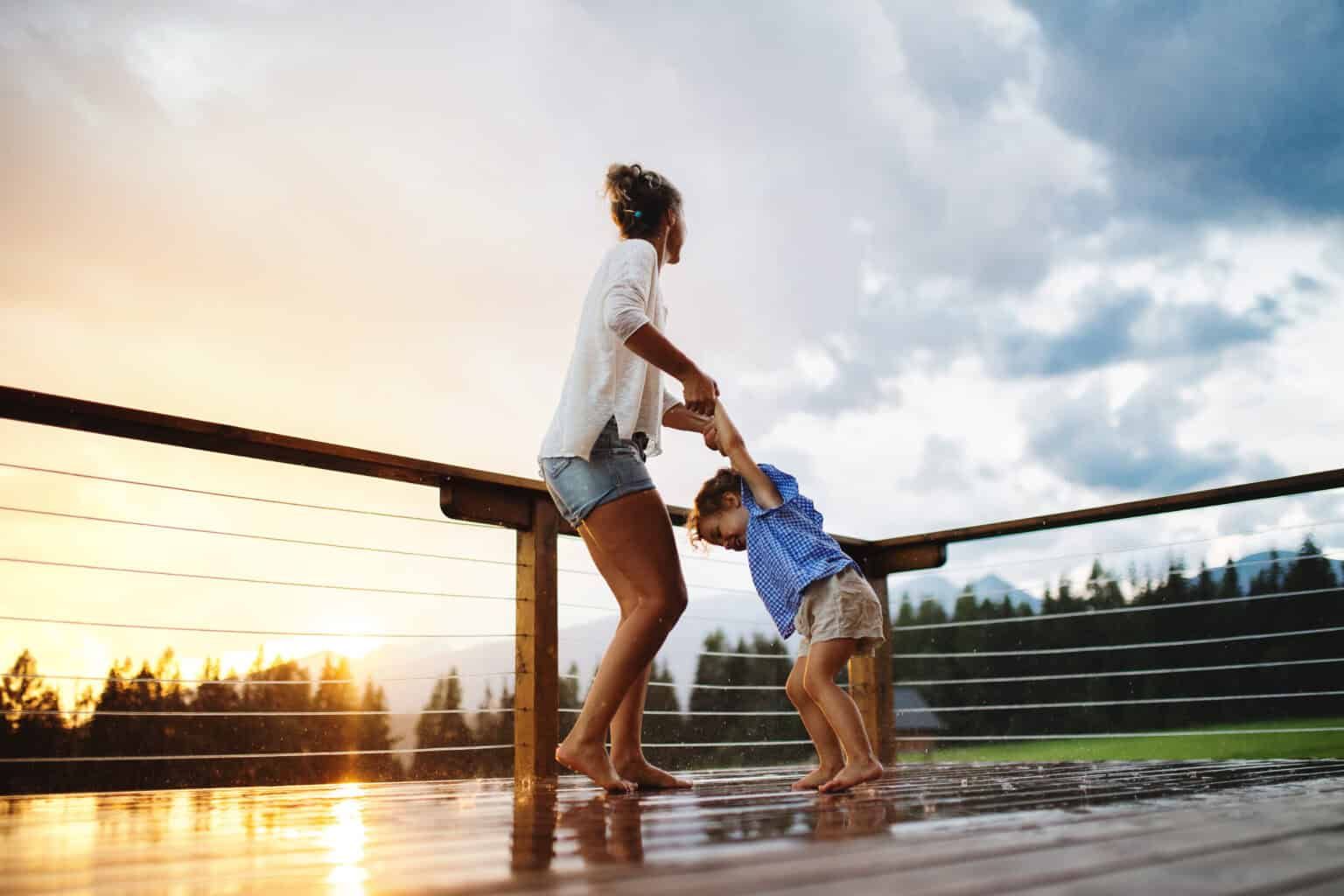 Woman and child playing on a wooden deck, holding hands, with mountains and sunset in background.