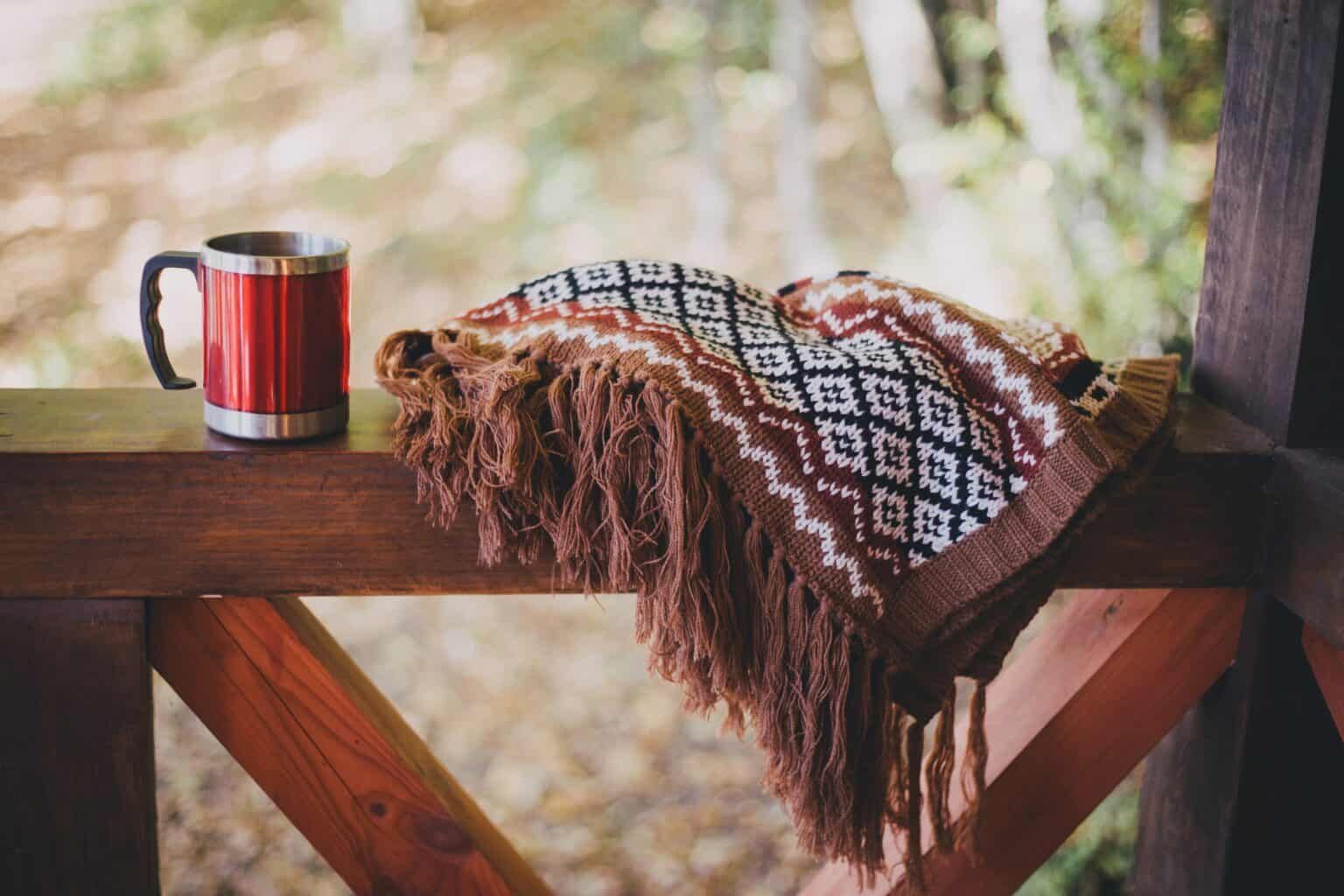 Red mug and patterned blanket on a wooden deck, blurred forest background.
