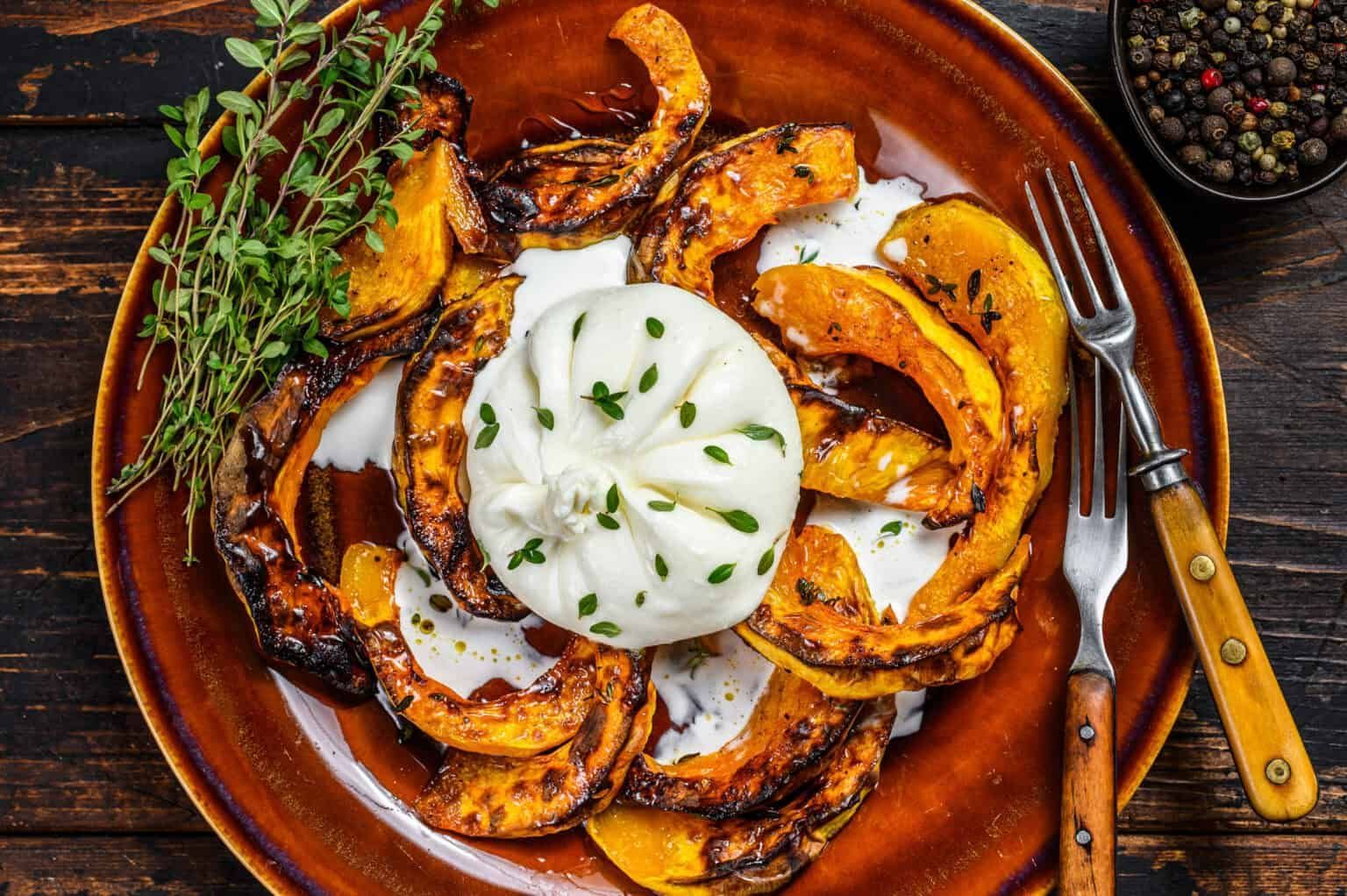 Roasted pumpkin slices with creamy cheese on a brown plate, garnished with herbs and a pepper grinder.