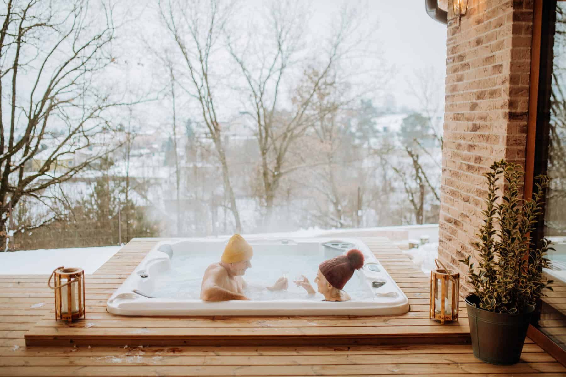 Two people in hats enjoy a hot tub on a snowy deck, overlooking a winter landscape.