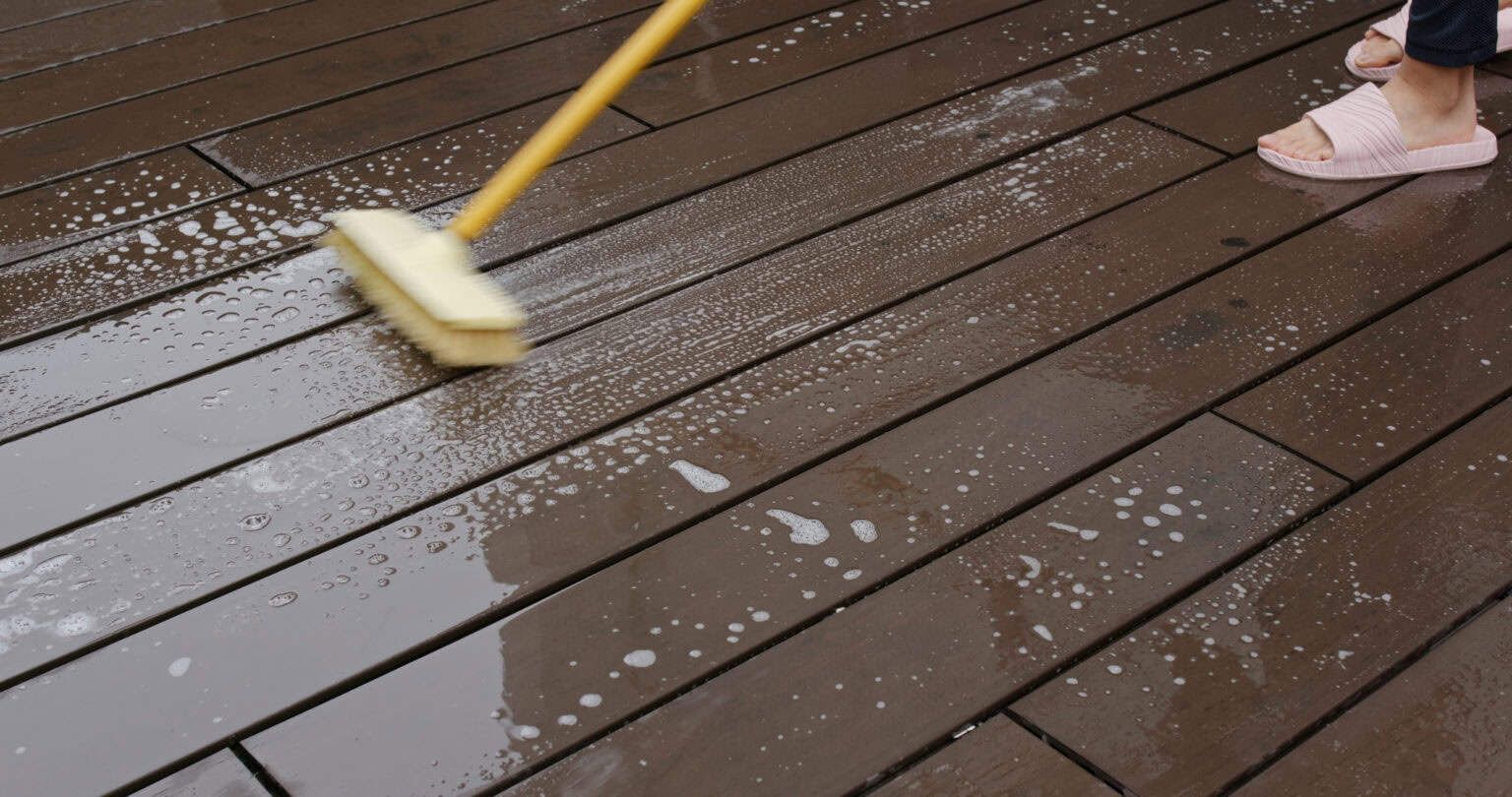 Person cleaning a wooden deck with a brush and soapy water.