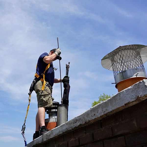 A technician in a safety harness cleans a chimney from a brick rooftop under a clear blue sky.