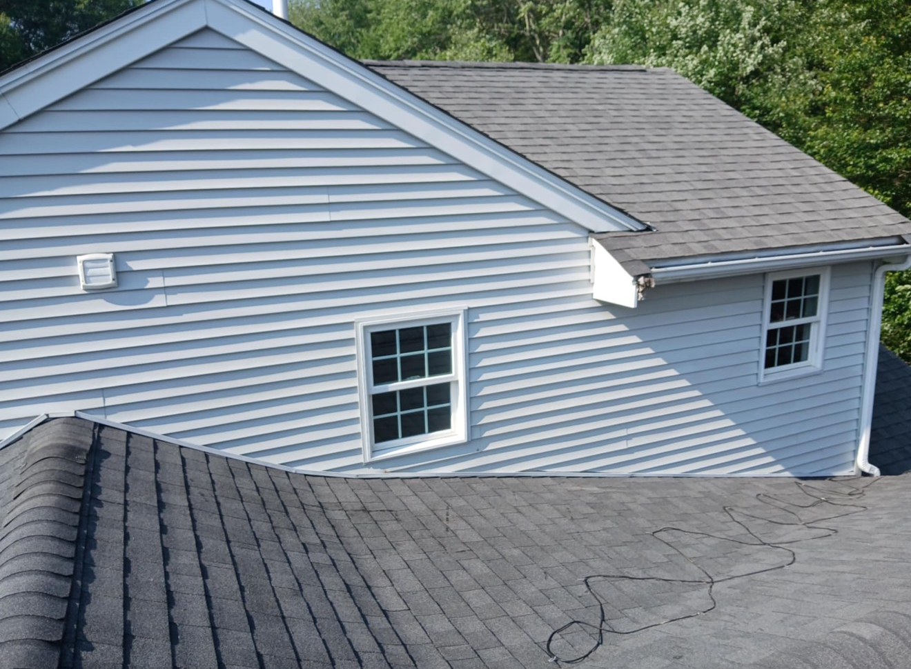 House with light blue siding, gray shingle roof, white windows, and trees in the background.