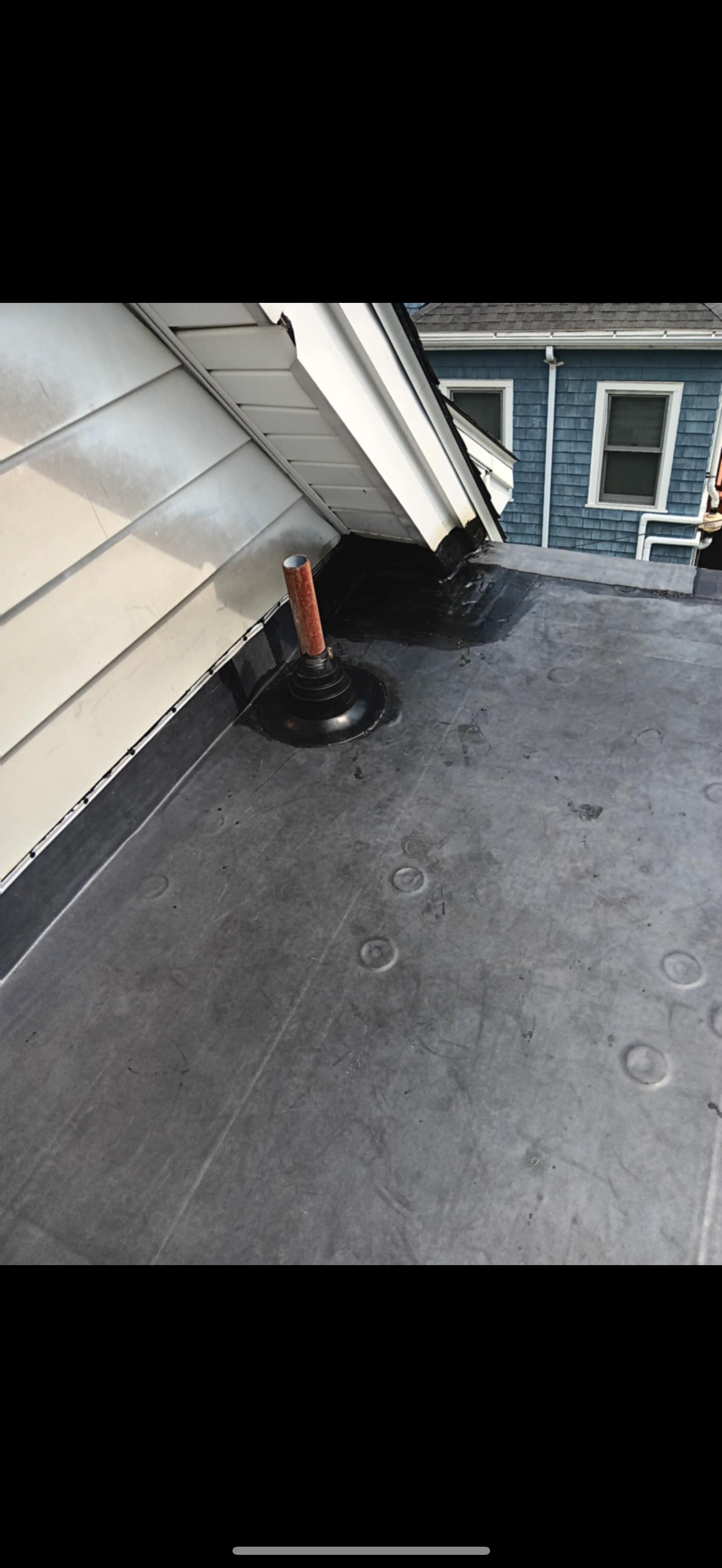 A black, flat roof with vent pipes and a house in the background.