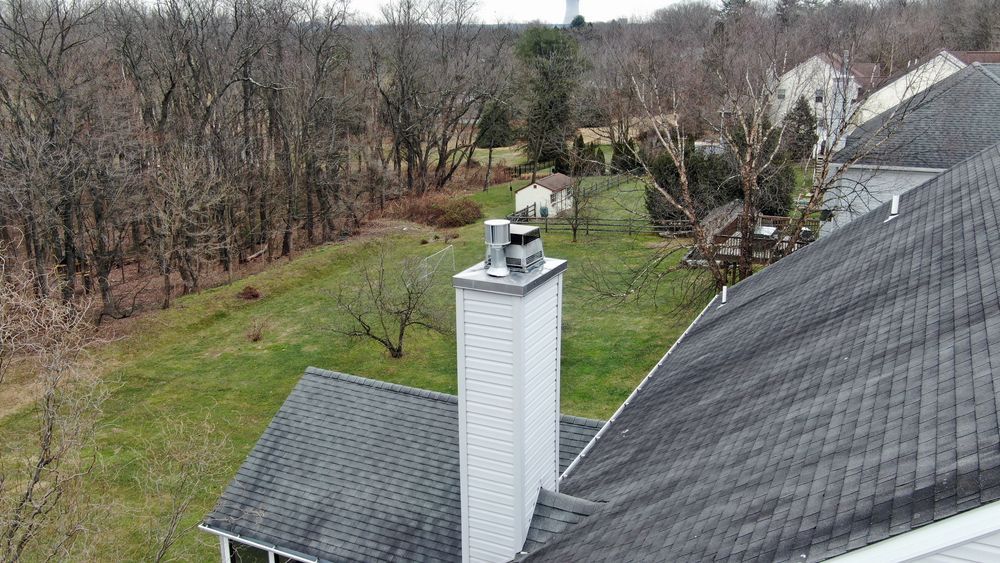 A gray rooftop with a chimney, overlooking a backyard with trees, a shed, and lawn.