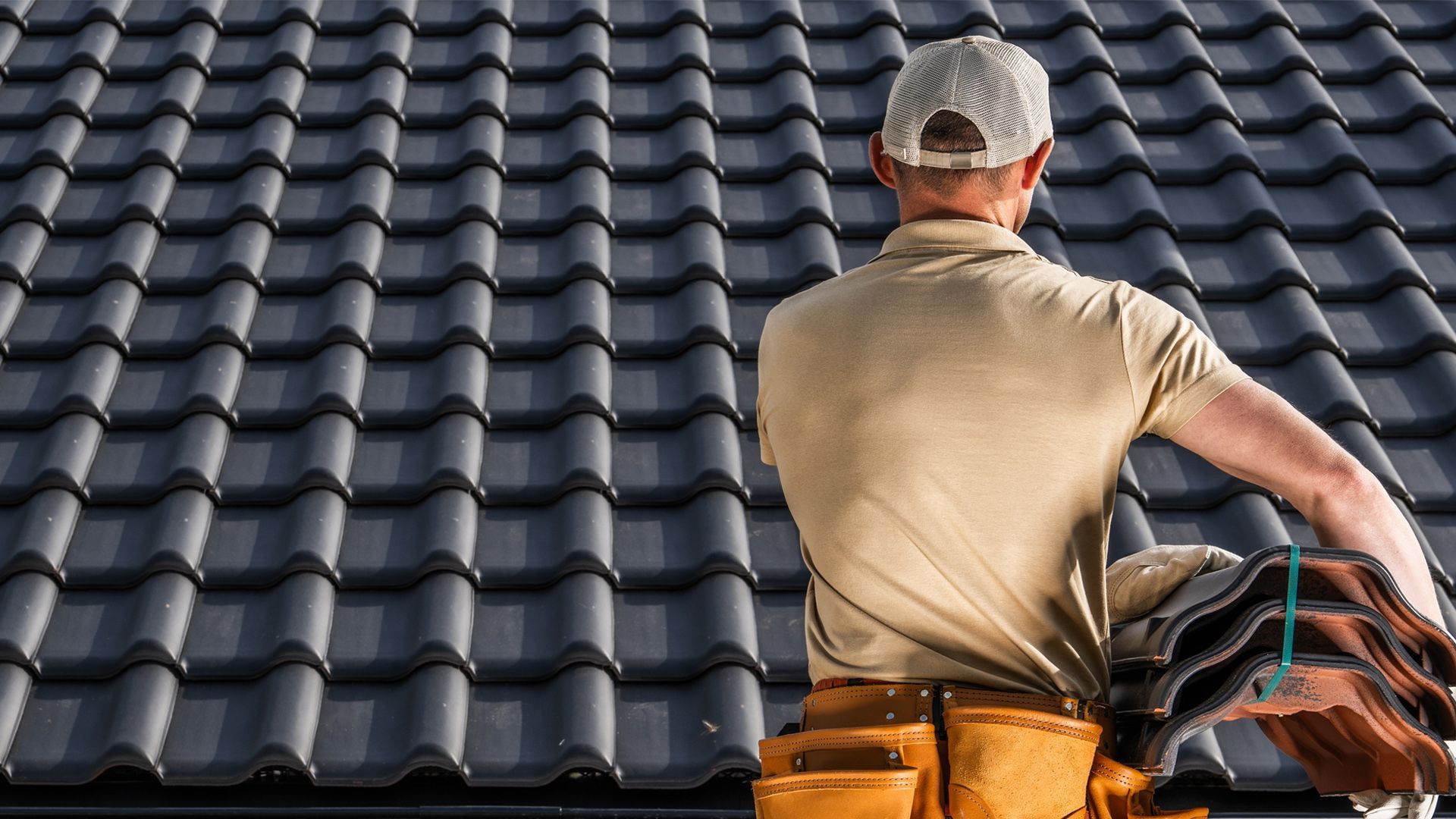 Roofer, wearing a cap and tool belt, holds roofing tiles while standing on a dark gray tiled roof.