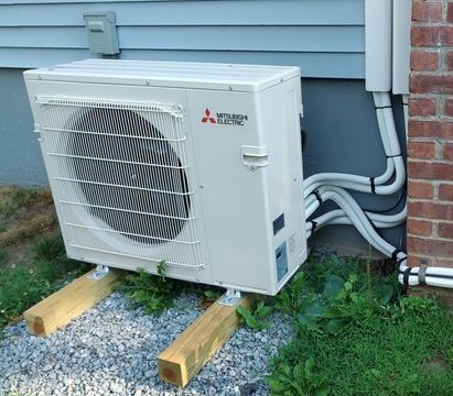 Mitsubishi Electric heat pump unit, supported by wooden beams on gravel, mounted next to a house with electrical conduits.
