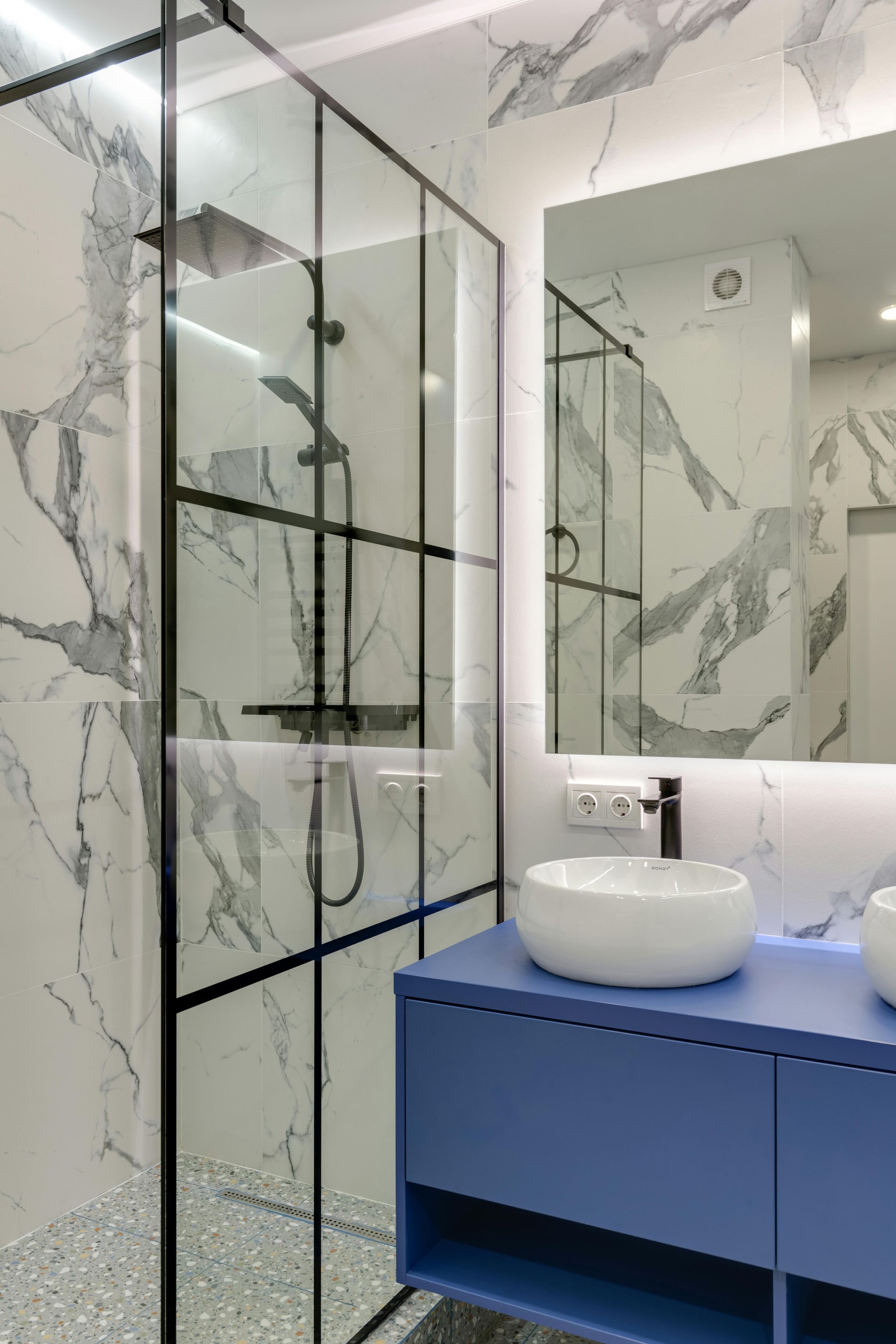 Modern bathroom with blue vanity, marble-patterned walls, and a black-framed glass shower. White vessel sink and large mirror.