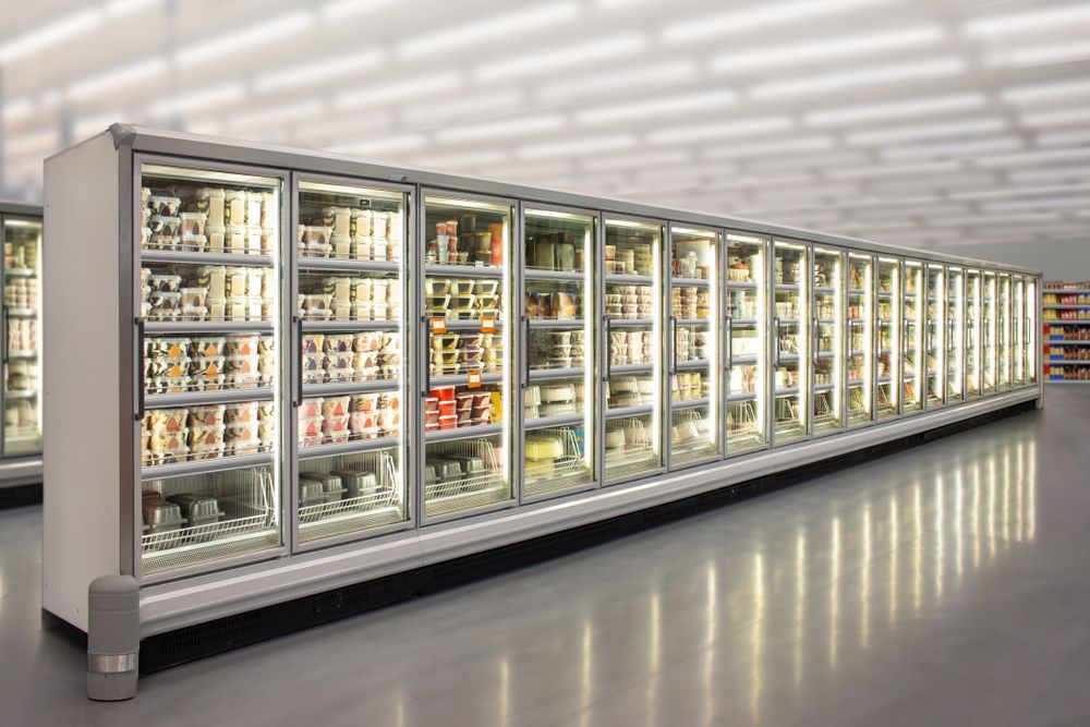 A long row of glass-door refrigerators in a brightly lit grocery store, filled with various food items.