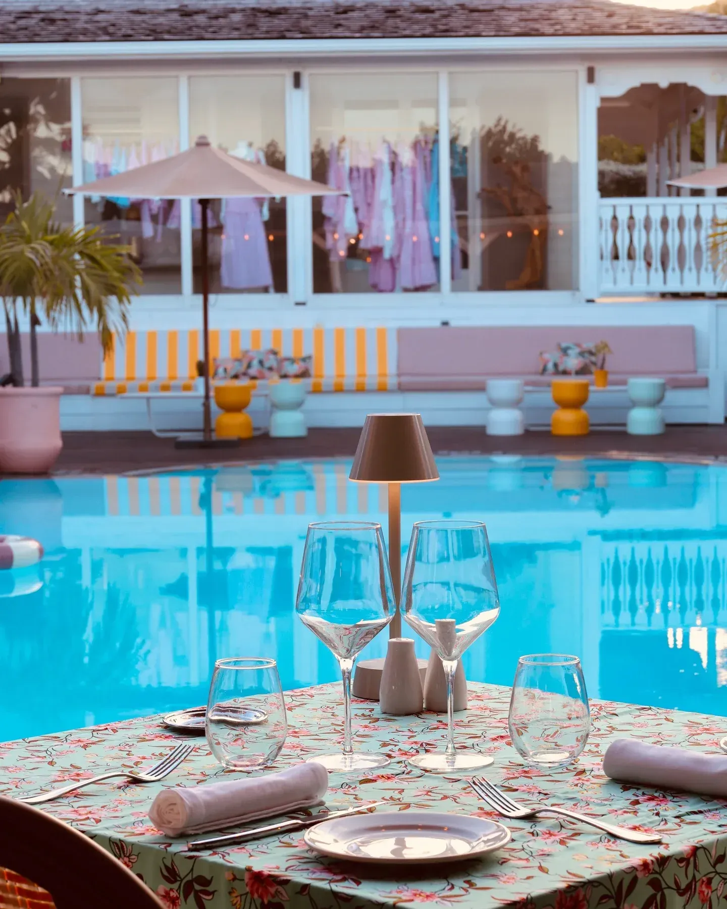 Table set for dinner by a pool, with glassware, floral tablecloth, and a lamp. The background shows a building with clothing and an umbrella.