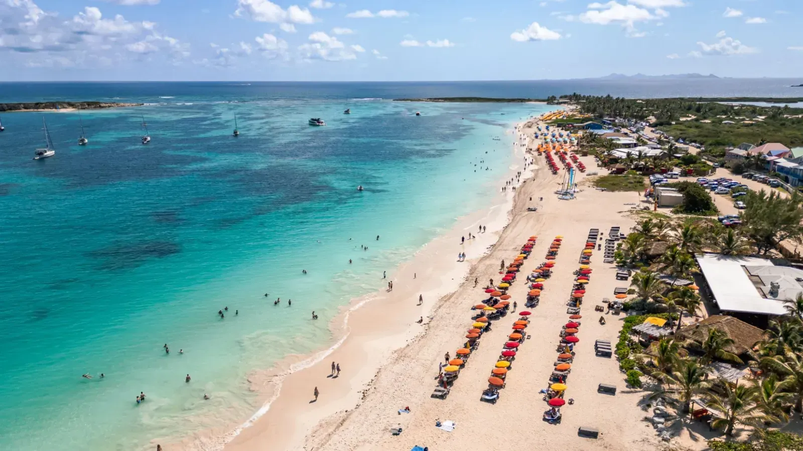 Aerial view of a sunny beach with turquoise water, lined with colorful umbrellas and lounge chairs, people swimming.
