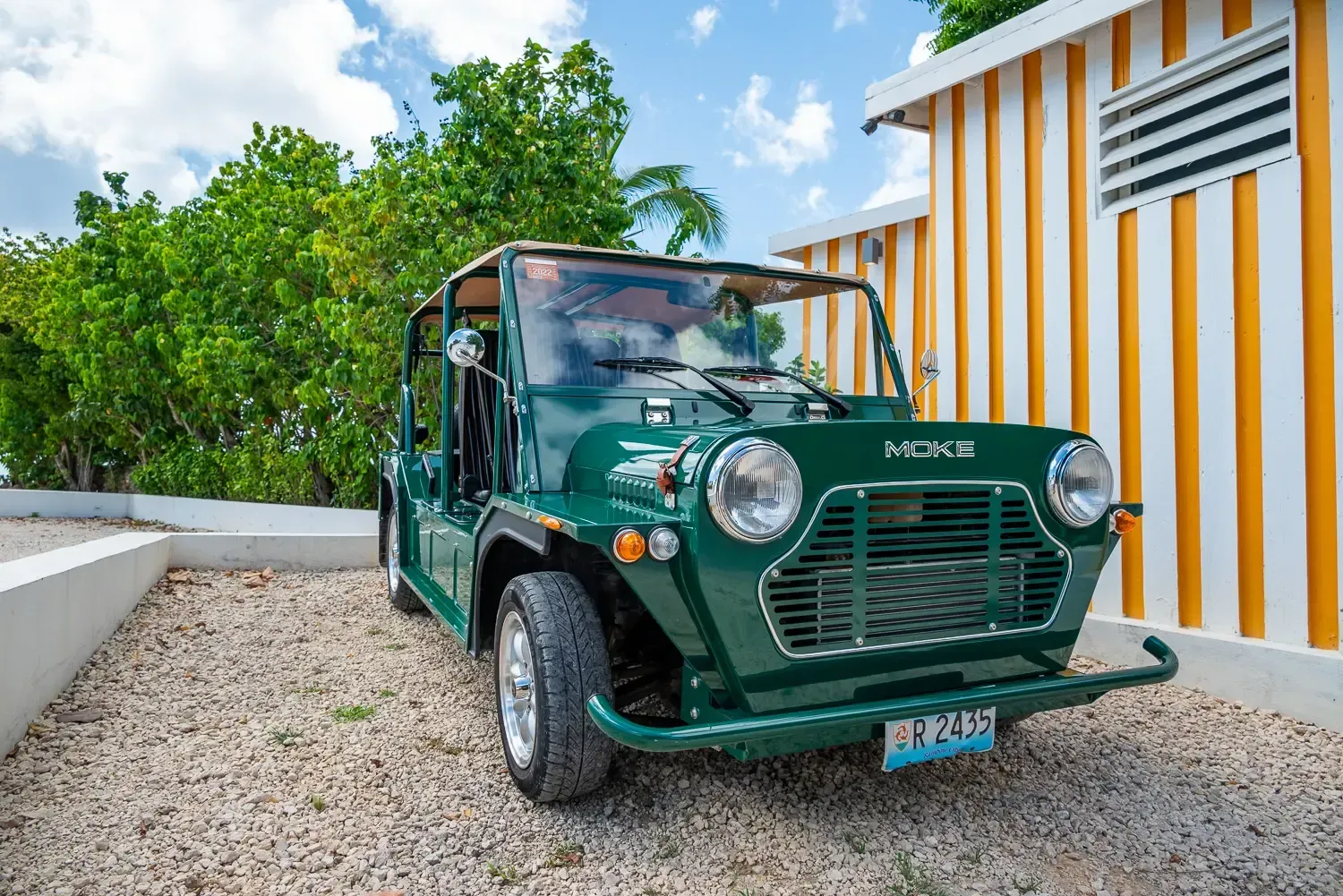 Green vintage Mini Moke car parked on gravel, beside a yellow and white striped building.