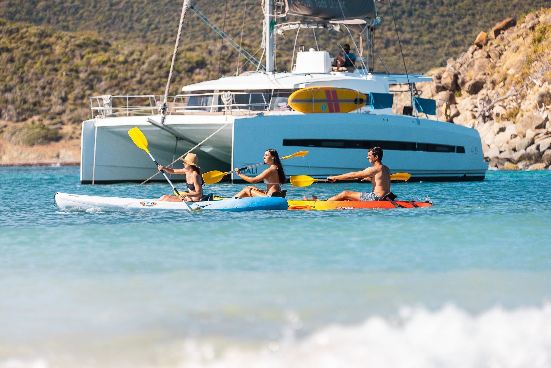 People kayaking near a catamaran in turquoise water; mountain backdrop.