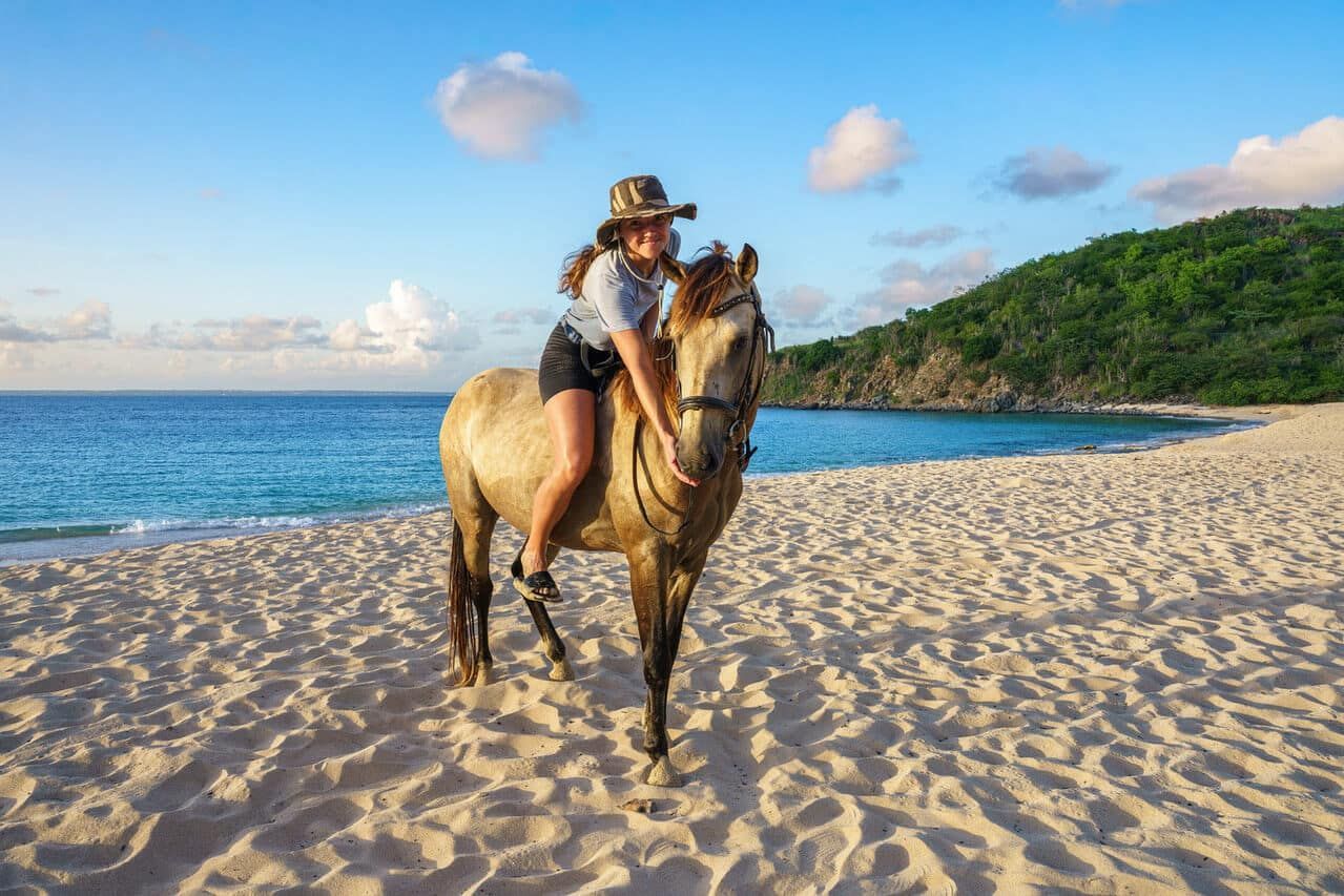 Woman riding a horse on a sandy beach. Turquoise water, green hills, and blue sky in the background.