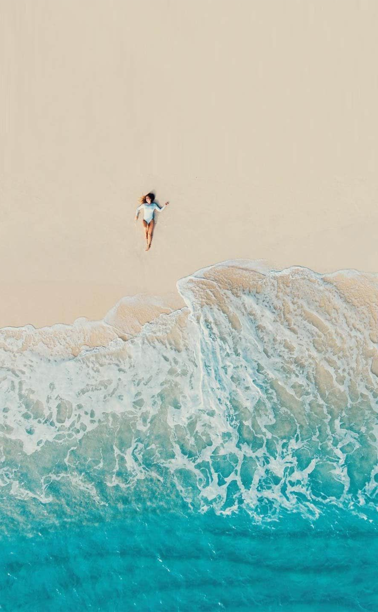 Woman lies on sandy beach as turquoise waves crash.