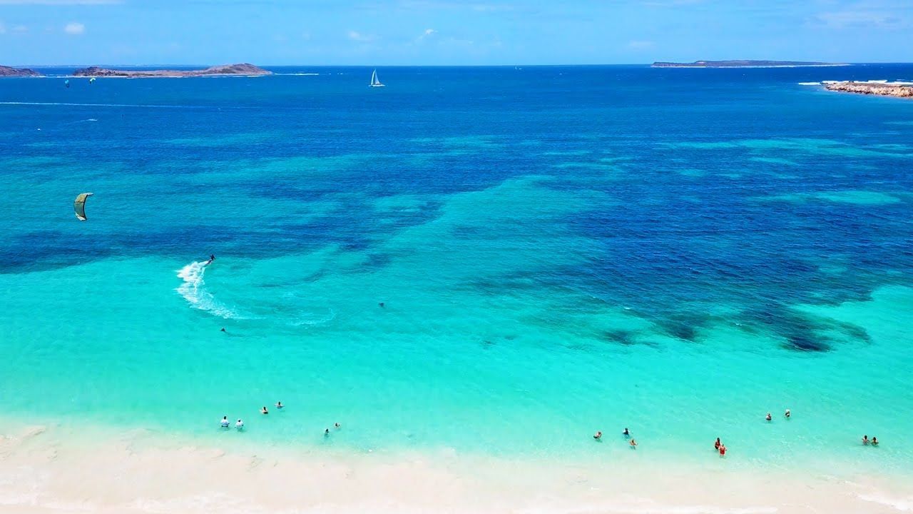 Bright turquoise ocean with white sand beach and people swimming. Blue sky, distant islands.