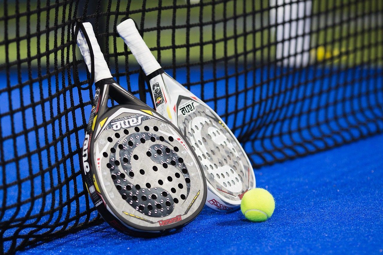 Two padel rackets and a yellow ball resting on a blue court next to a black net.