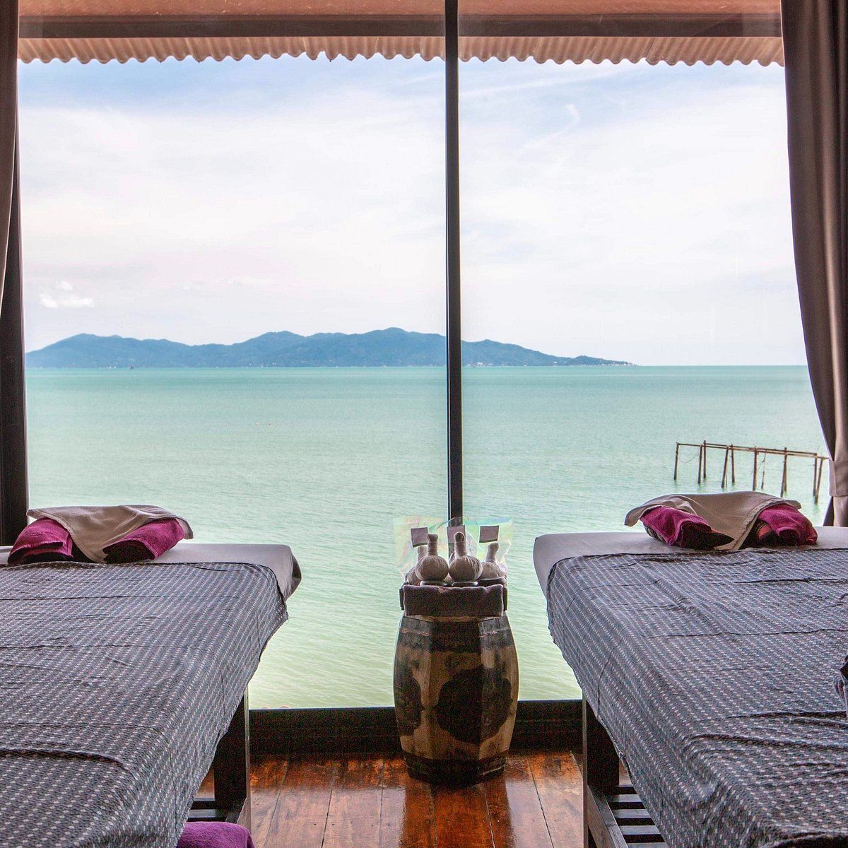 Spa treatment room with ocean view, two massage tables, towels, and a decorative jar.
