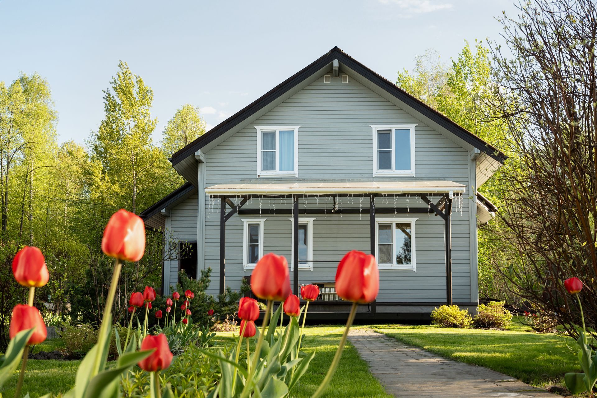 Gray two-story house with a porch, red tulips in the foreground, and trees in the background.