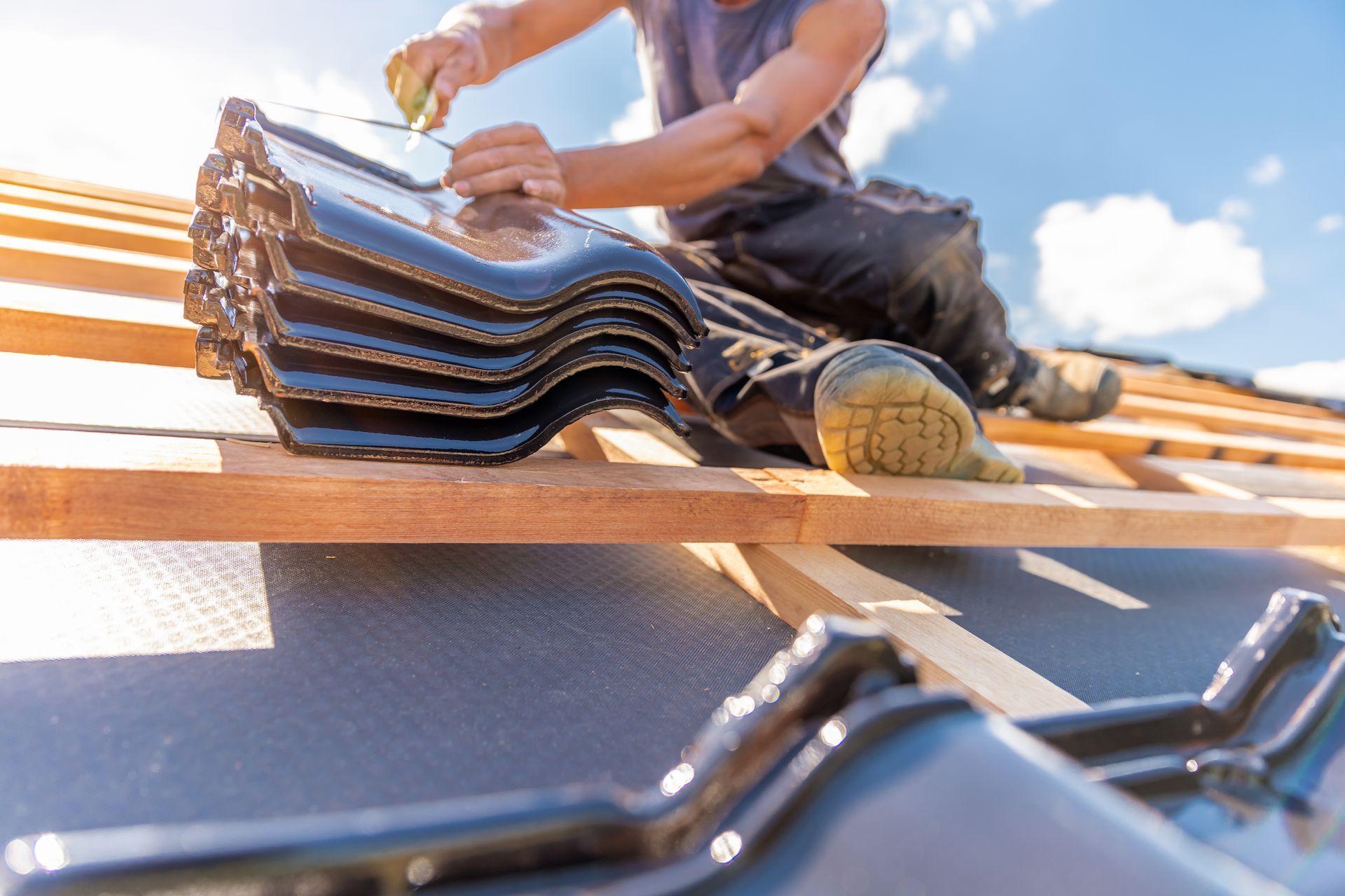 Roofer installing dark tiles on a wooden roof frame under a blue sky.