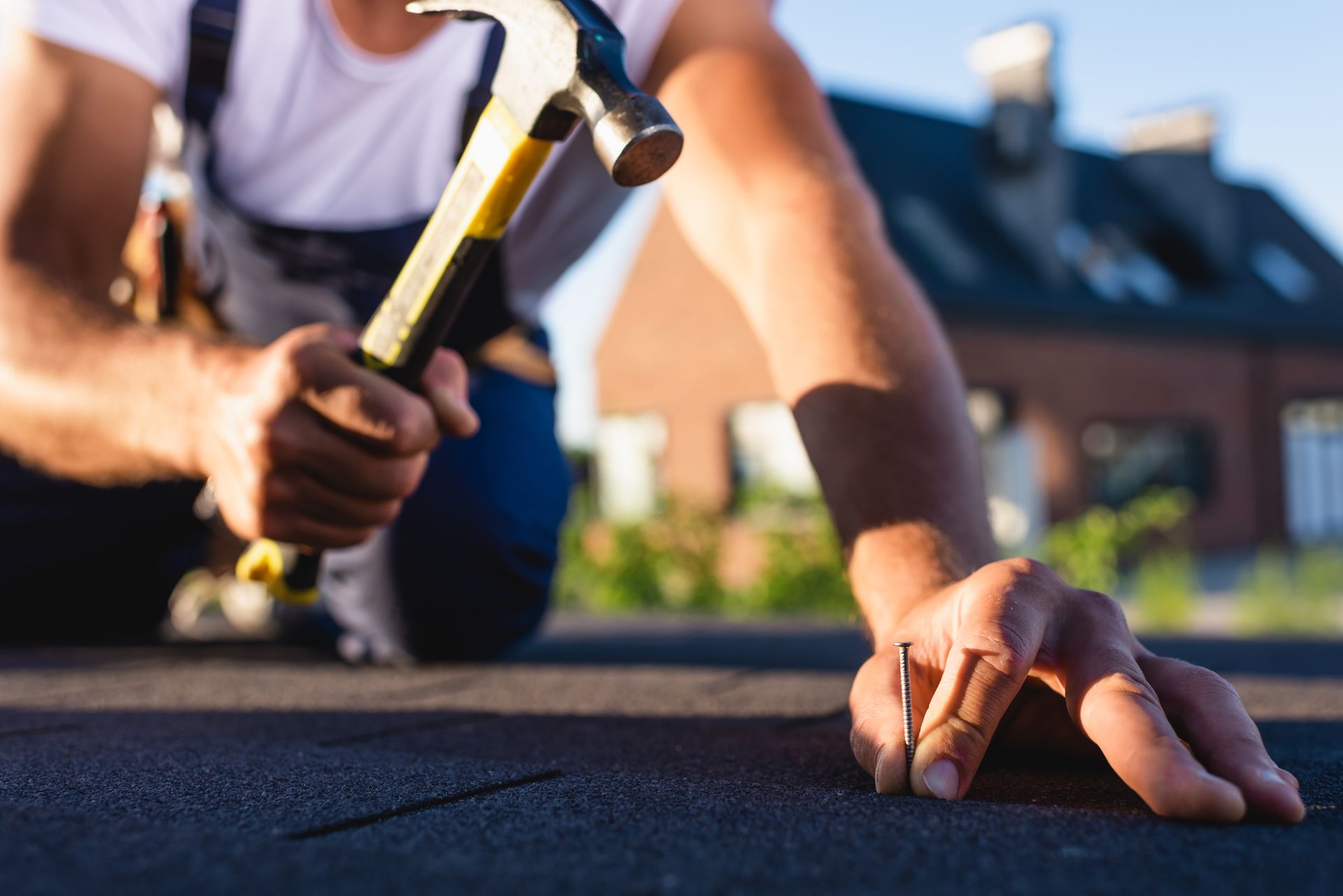 Roofer hammering a nail into dark roofing material, in front of a house.