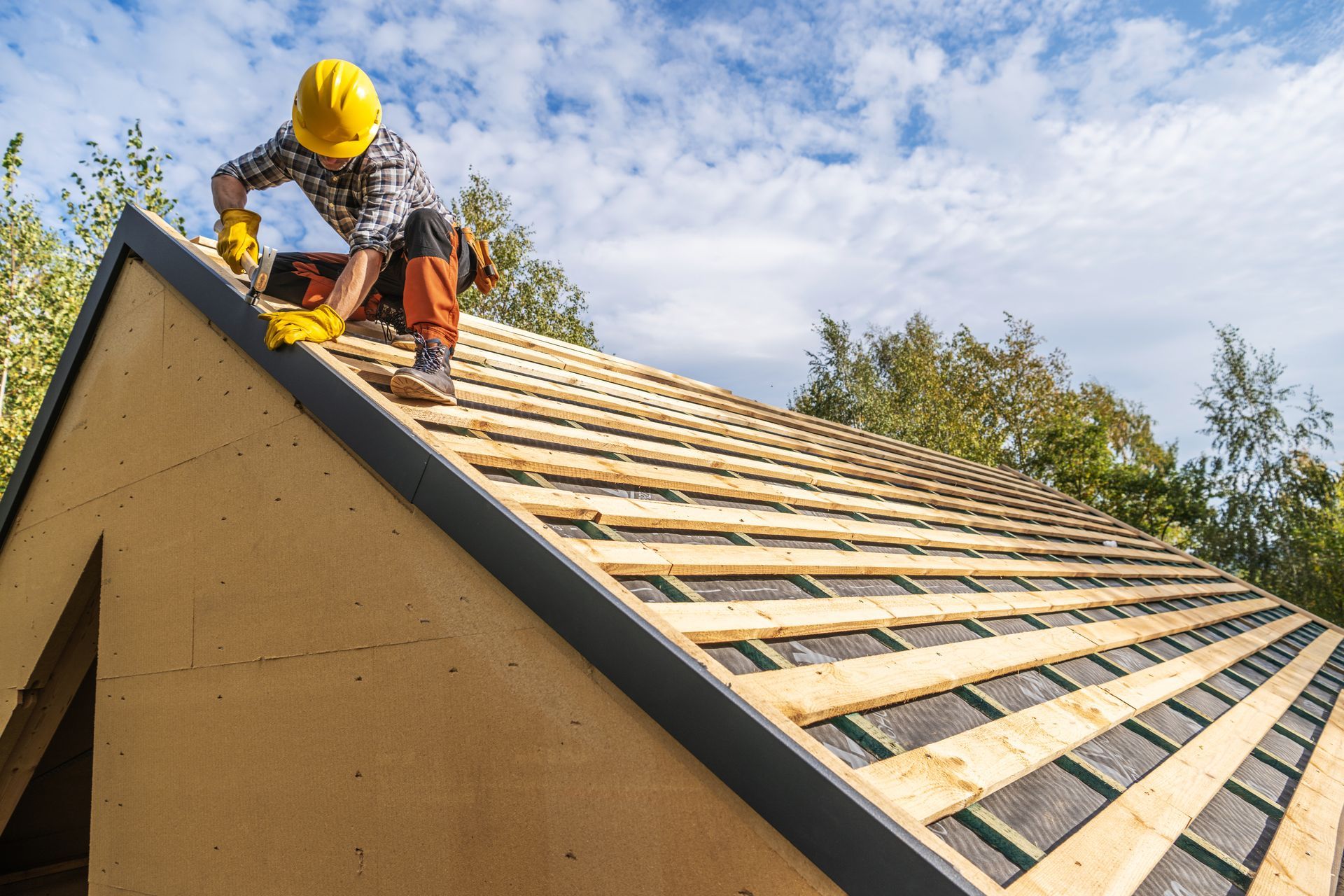 A roofer in a yellow helmet installs shingles on a wooden roof frame under a blue sky.