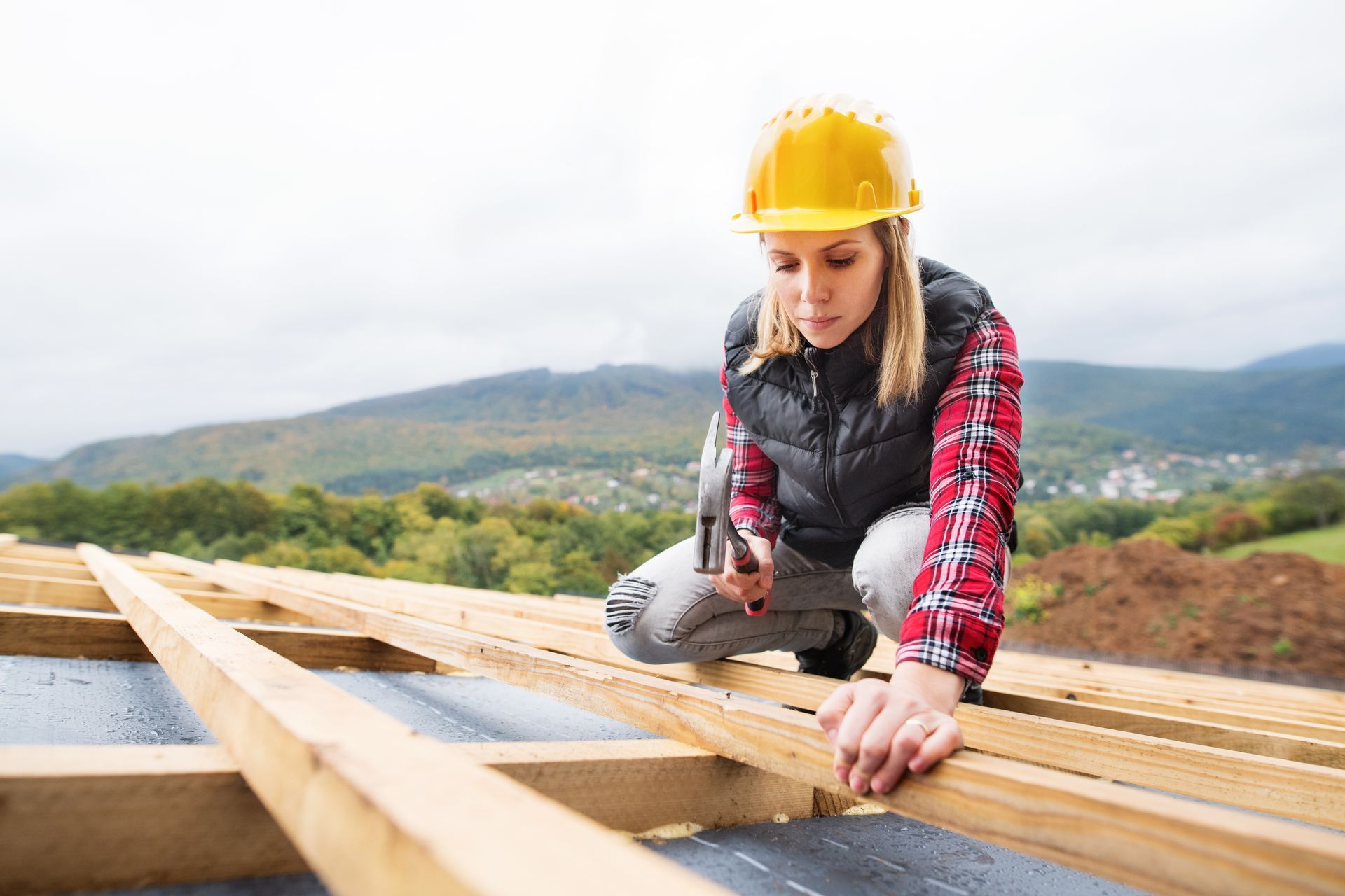 Woman in yellow hard hat hammers wood on a roof, mountains in the background.