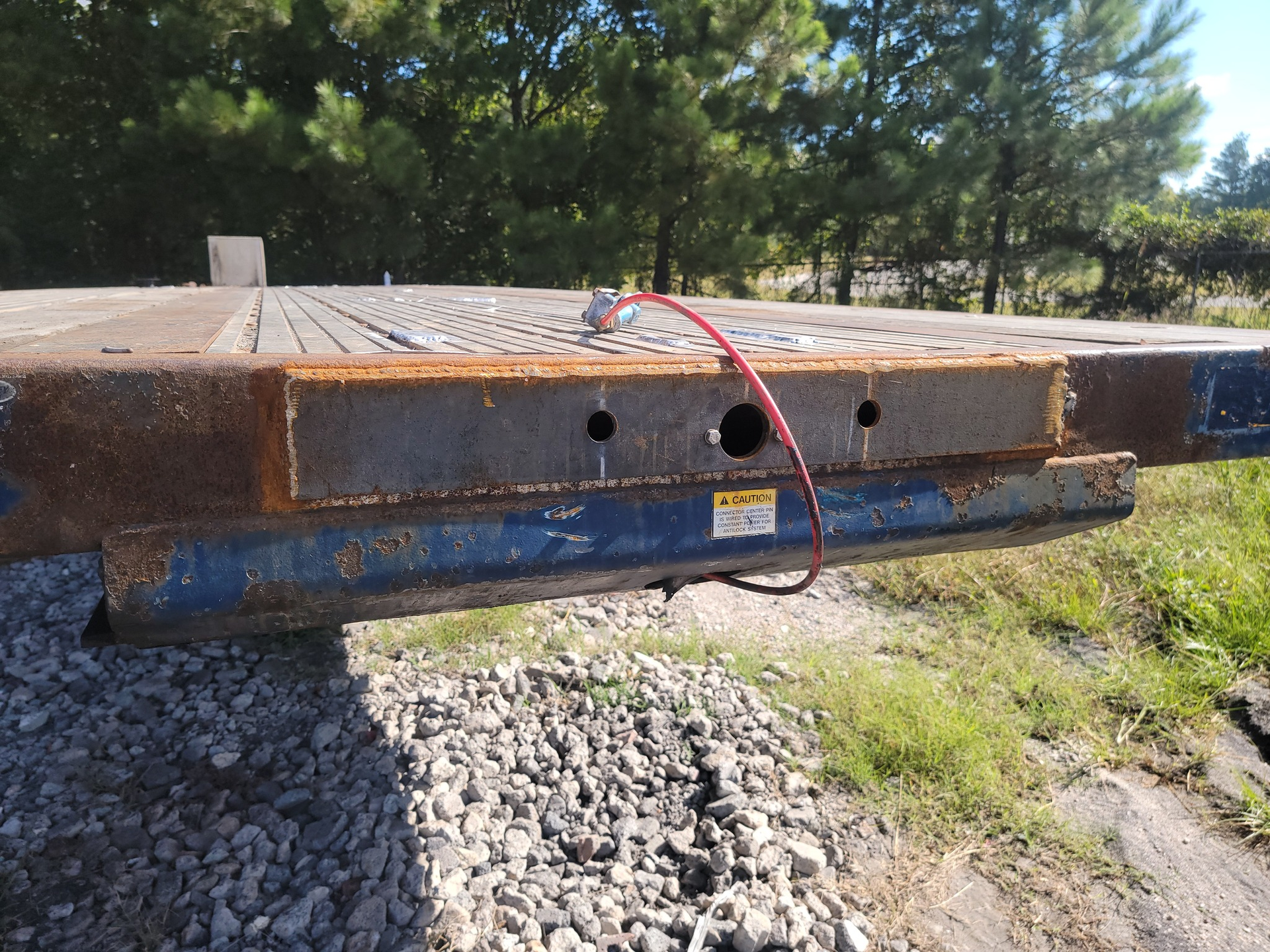 Close-up of a flatbed trailer's front end, showing steel construction, rust, and a red coiled wire against a backdrop of trees.